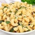 A bowl of cooked cavatappi pasta mixed with slices of zucchini, topped with grated cheese, chopped basil, and cracked black pepper.
