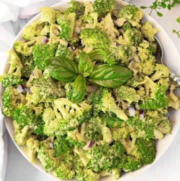 A bowl of fresh broccoli salad with chopped red onions, creamy dressing, and a garnish of basil leaves on top. A spoon is placed in the bowl.