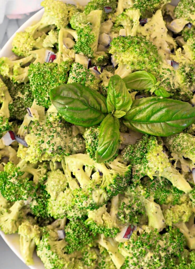 A close-up of a broccoli salad topped with a creamy dressing and garnished with a cluster of fresh basil leaves in the center.