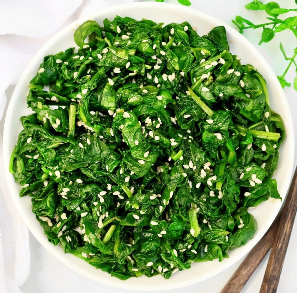 A white bowl filled with cooked spinach leaves, garnished with white sesame seeds. Wooden chopsticks and a white napkin are placed nearby on a white surface.