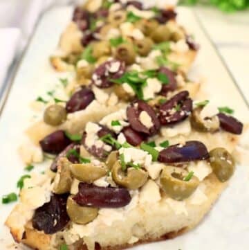 A close-up of toasted bread slices topped with crumbled feta cheese, chopped green and black olives, and garnished with fresh chopped herbs, arranged on a white rectangular plate.