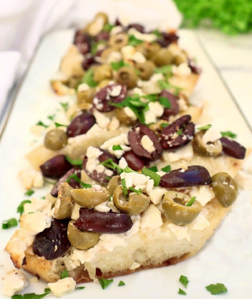 A close-up of toasted bread slices topped with crumbled feta cheese, chopped green and black olives, and garnished with fresh chopped herbs, arranged on a white rectangular plate.