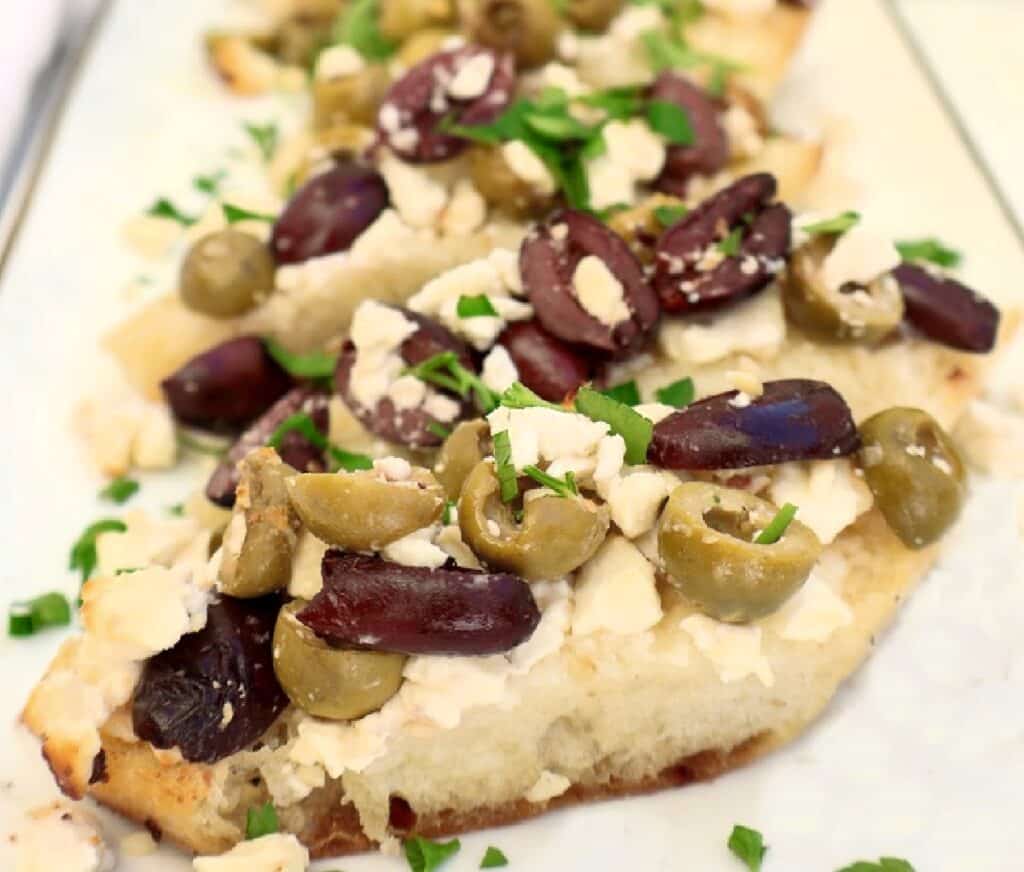 Slices of flatbread topped with chopped green and black olives, crumbled feta cheese, and fresh herbs, arranged on a white serving tray.