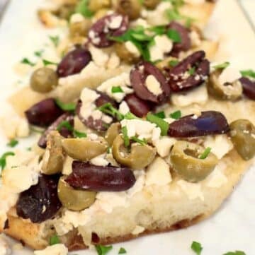 Slices of flatbread topped with chopped green and black olives, crumbled feta cheese, and fresh herbs, arranged on a white serving tray.