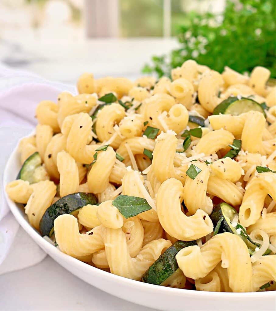 A close-up of a white bowl filled with curly pasta, zucchini slices, grated cheese, and fresh basil, garnished with black pepper. Green herbs are visible in the background.