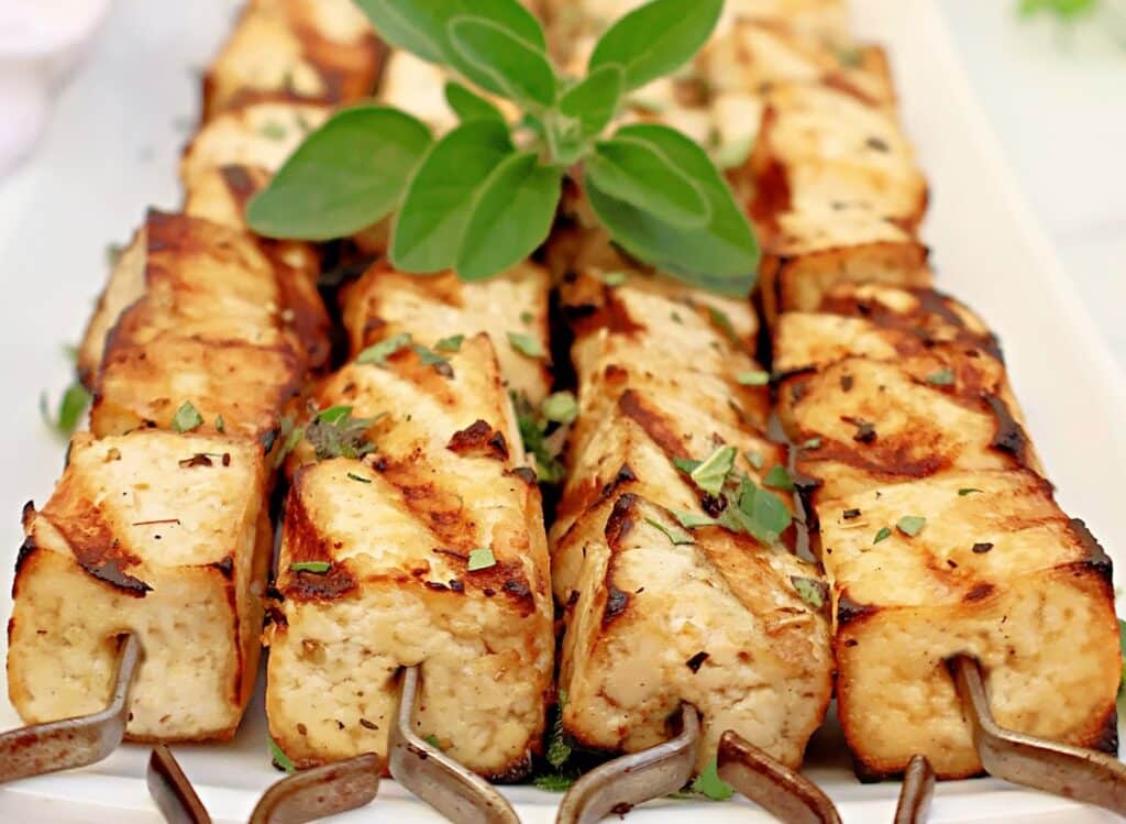 Grilled tofu skewers with golden brown edges, garnished with fresh herbs and a sprig of green on top, served on a white platter.