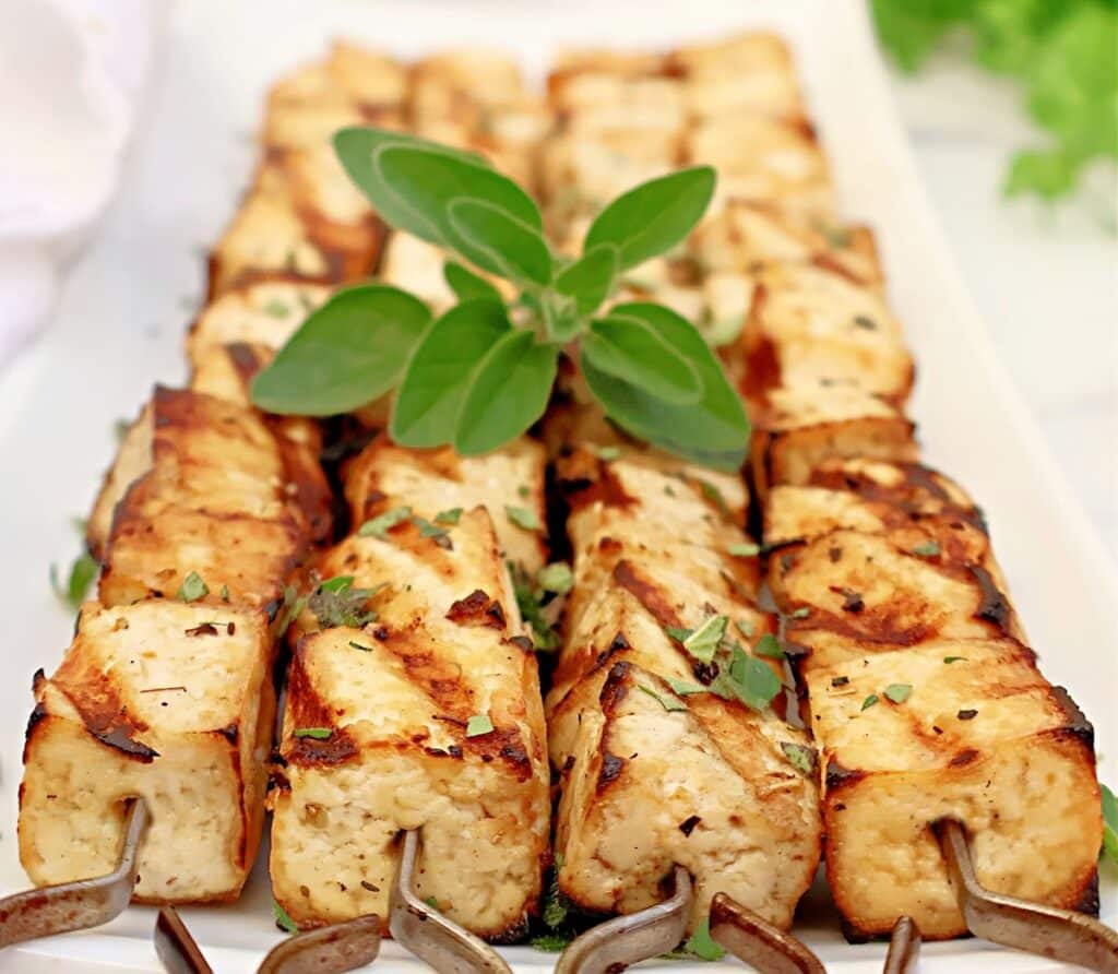 Grilled tofu skewers arranged neatly on a rectangular white plate, garnished with fresh herbs and a sprig of green leaves on top.