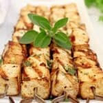Grilled tofu skewers arranged neatly on a rectangular white plate, garnished with fresh herbs and a sprig of green leaves on top.