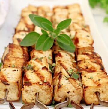 Grilled tofu skewers arranged neatly on a rectangular white plate, garnished with fresh herbs and a sprig of green leaves on top.