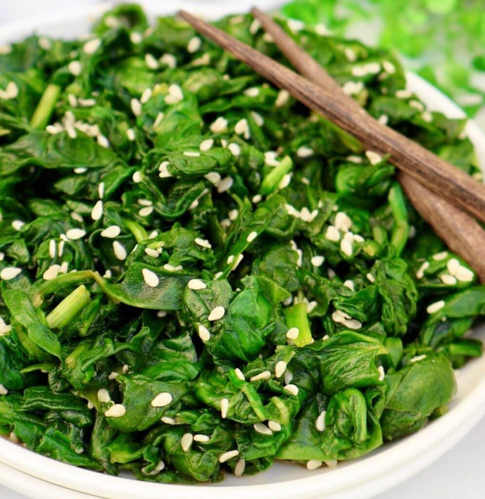 A plate of sautéed spinach garnished with white sesame seeds, with a pair of wooden chopsticks resting on the edge.