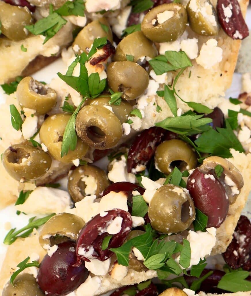 Close-up of focaccia bread topped with green and kalamata olives, crumbled feta cheese, and fresh chopped parsley.