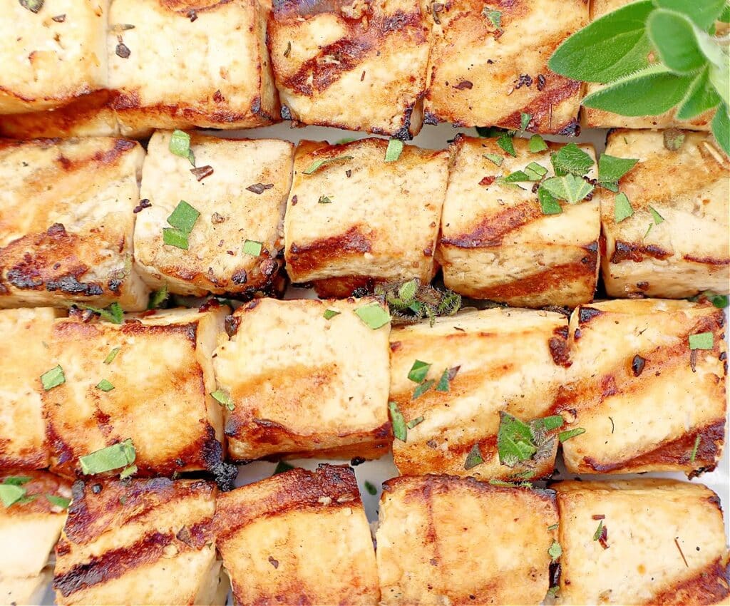 Grilled tofu skewers with golden-brown char marks, garnished with chopped fresh herbs, arranged closely together.