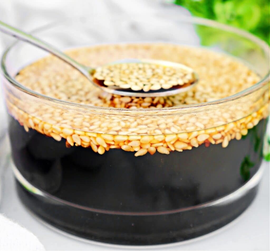 A glass bowl filled with dark soy sauce topped with floating sesame seeds, with a spoon lifting some seeds from the surface.