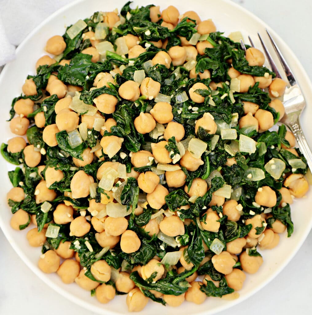A plate of sautéed spinach, chickpeas, and diced onions is served with a fork on the side. The dish appears light and healthy, featuring green and beige colors.