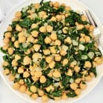 A plate of sautéed spinach, chickpeas, and diced onions is served with a fork on the side. The dish appears light and healthy, featuring green and beige colors.