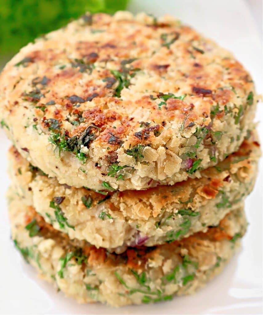 Three golden-brown, herb-filled patties are stacked on a white plate, showing a crispy, textured surface and visible flecks of green herbs throughout.
