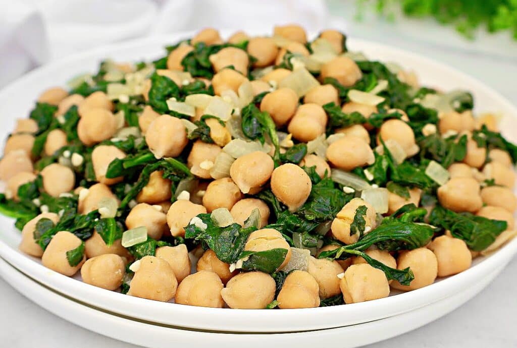 A white plate filled with a chickpea and spinach salad, mixed with cooked onions and garlic. The dish appears fresh and healthy, served on a white surface with a blurred green background.