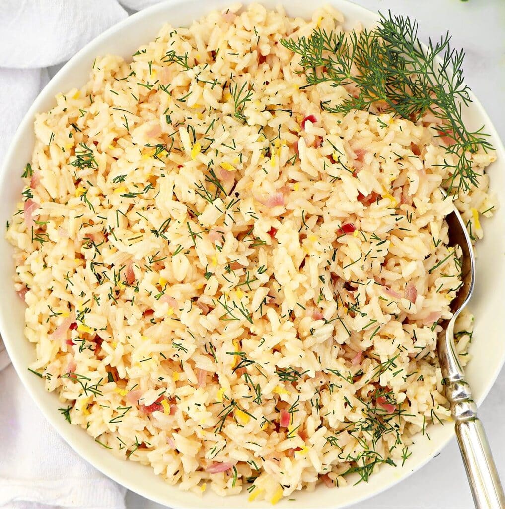 A white bowl filled with cooked rice garnished with fresh dill, with a silver spoon resting on the side of the bowl. The rice appears fluffy and mixed with small bits of onion or herbs.