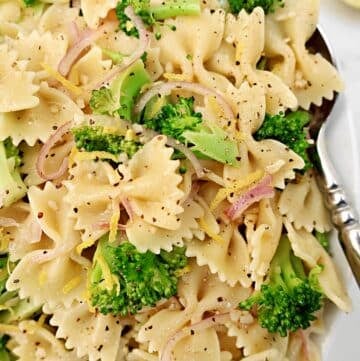 Close-up of a plate of bow tie pasta mixed with broccoli florets, thinly sliced red onions, and black pepper, garnished with lemon zest. A silver fork and lemon slices are visible on the side.