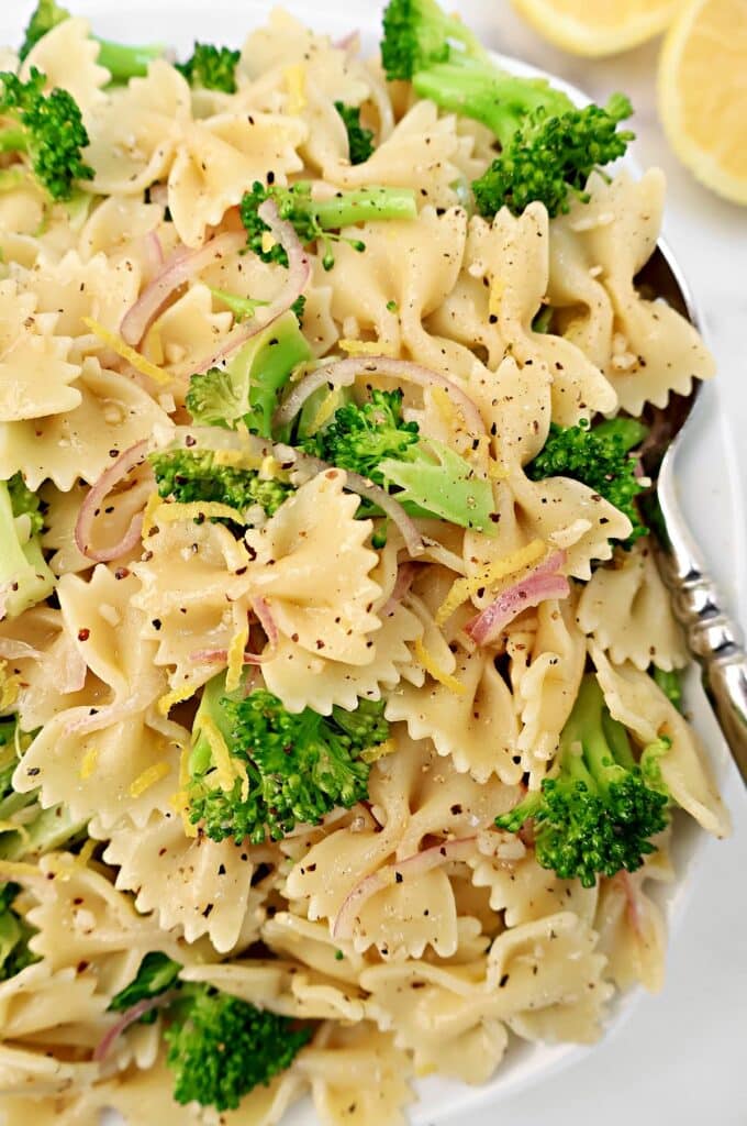 Close-up of a plate of bow tie pasta mixed with broccoli florets, thinly sliced red onions, and black pepper, garnished with lemon zest. A silver fork and lemon slices are visible on the side.