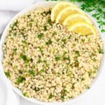 A bowl of cooked pearl couscous garnished with chopped parsley, with four lemon slices arranged on the side. The dish is presented on a white background with a sprig of greenery in the corner.