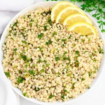 A bowl of cooked pearl couscous garnished with chopped parsley, with four lemon slices arranged on the side. The dish is presented on a white background with a sprig of greenery in the corner.