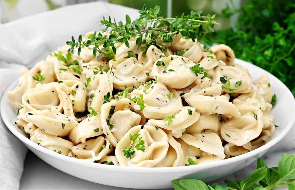 A white bowl filled with cooked tortellini pasta, garnished with chopped fresh herbs and cracked black pepper, sits on a table with green leaves and herbs in the background.