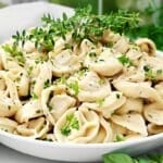 A white bowl filled with cooked tortellini pasta, garnished with chopped fresh herbs and cracked black pepper, sits on a table with green leaves and herbs in the background.
