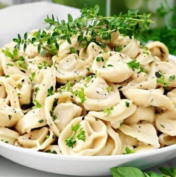 A white bowl filled with cooked tortellini pasta, garnished with chopped fresh herbs and cracked black pepper, sits on a table with green leaves and herbs in the background.