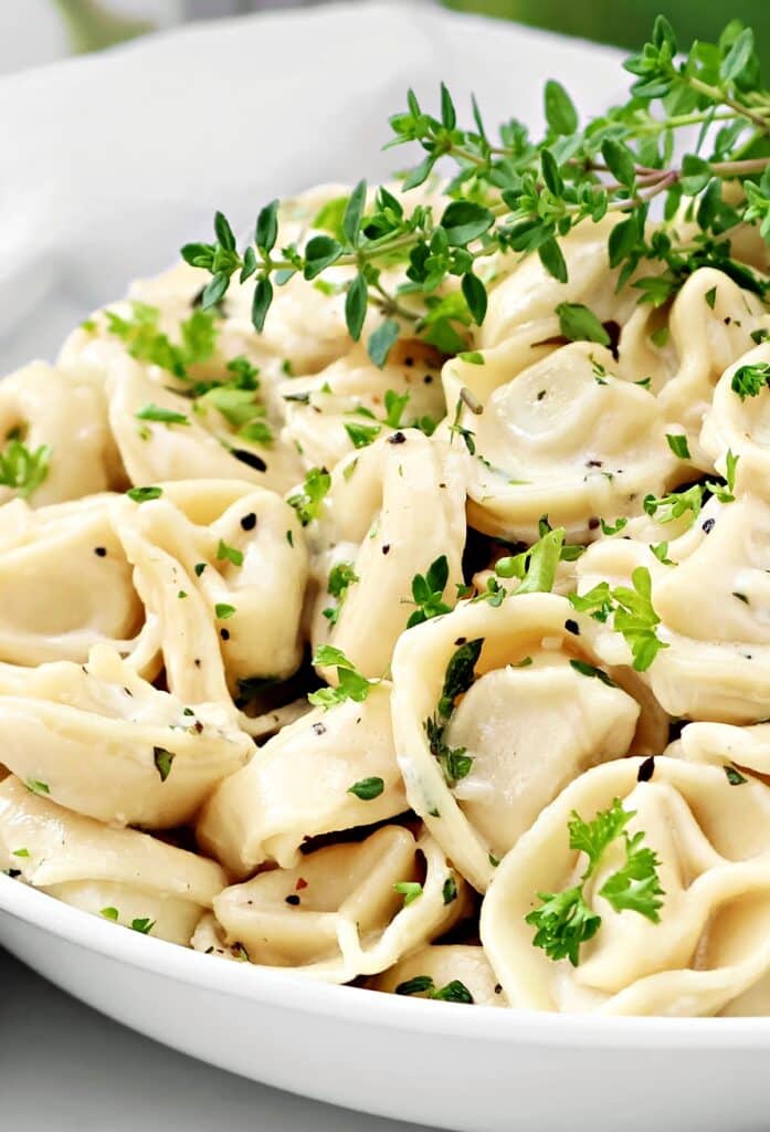A close-up of creamy tortellini pasta garnished with fresh green herbs, served in a white bowl.