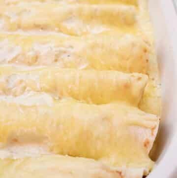 A close-up of a baking dish filled with several cheese-covered enciladas, topped with a creamy white sauce.