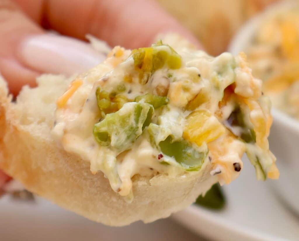 A close-up of a hand holding a piece of bread topped with a creamy dip containing chopped green peppers and shredded cheese.