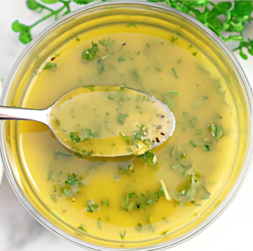 A spoonful of lemon vinaigrette with chopped green herbs is held over a bowl filled with the same dressing, with fresh herbs visible in the background.
