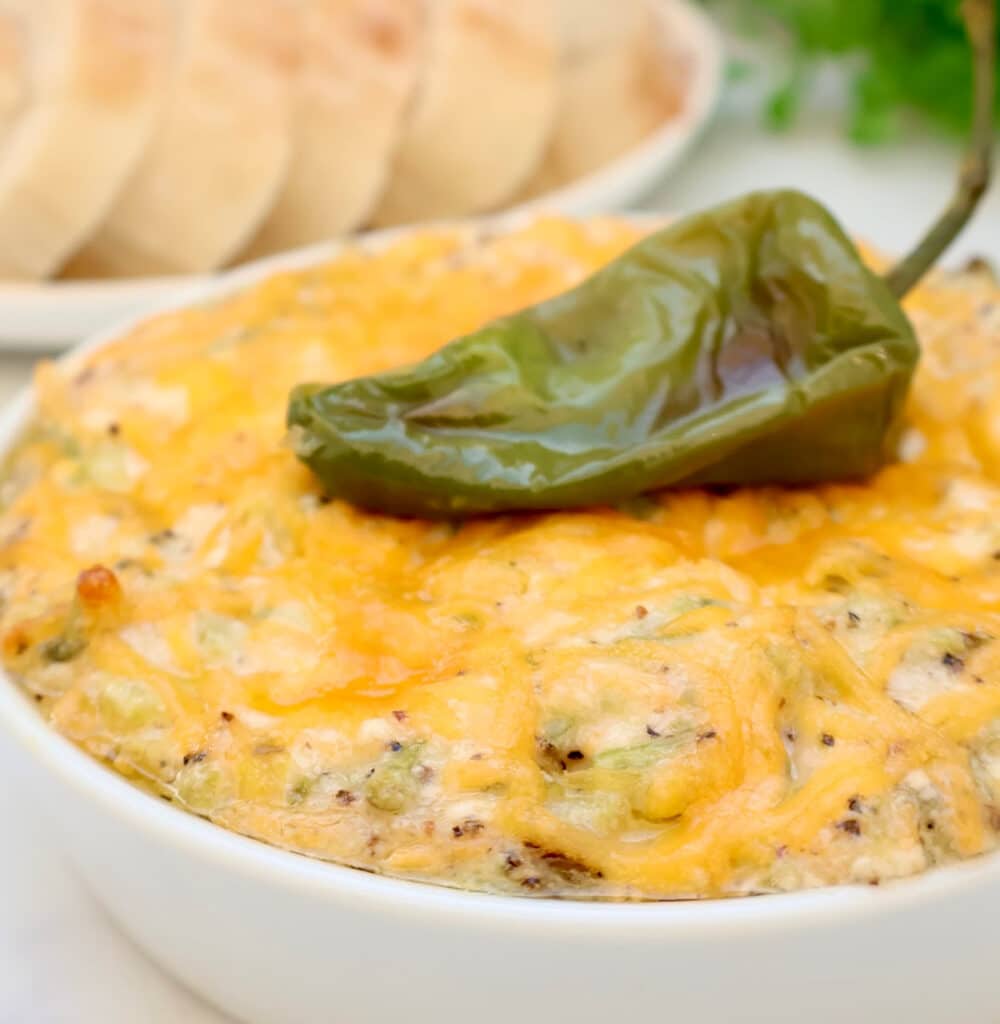 A bowl of cheesy, baked dip topped with a whole roasted green chili pepper, with a plate of sliced bread in the background.
