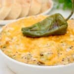 A bowl of cheesy, baked dip topped with a whole roasted green chili pepper, with a plate of sliced bread in the background.