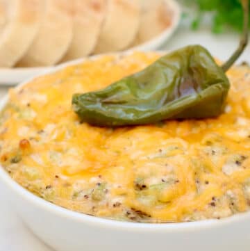 A bowl of cheesy, baked dip topped with a whole roasted green chili pepper, with a plate of sliced bread in the background.