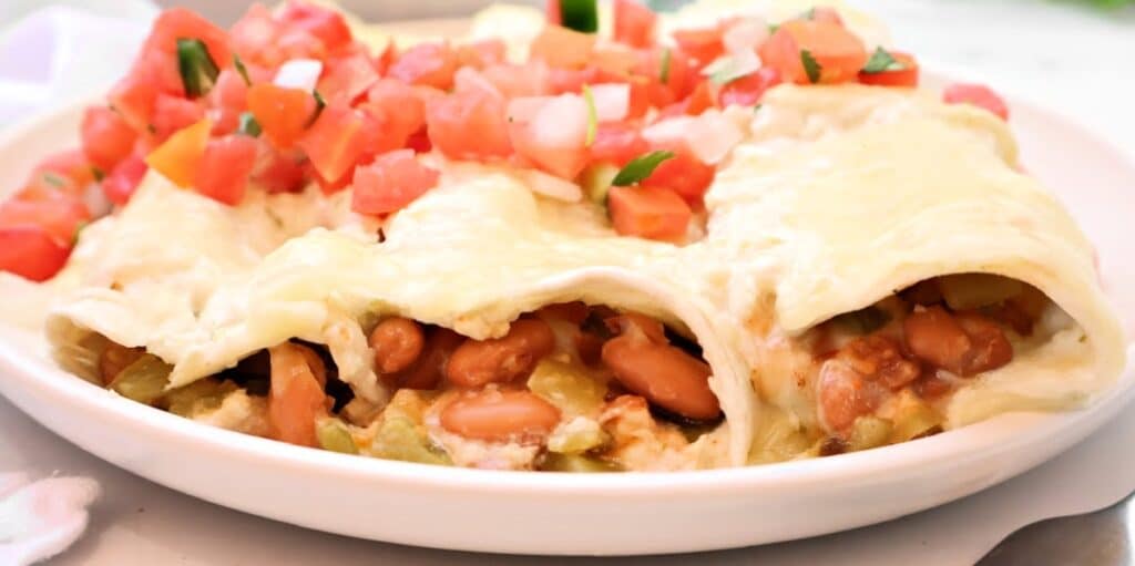 A close-up of cheesy enchiladas filled with beans and vegetables, topped with chopped tomatoes, onions, and jalapeños, served on a white plate.