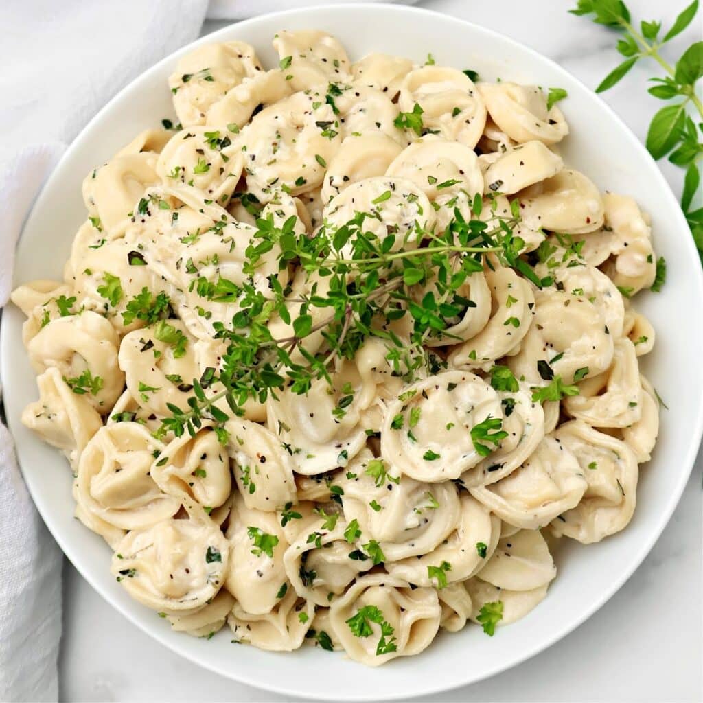 A white plate filled with creamy tortellini pasta, garnished with chopped parsley and a sprig of fresh herbs on top. The dish is set on a light surface with a napkin and some herbs nearby.