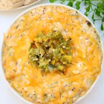 A bowl of baked cheesy dip topped with chopped green chilies sits on a white surface, with a sprig of greenery and a plate of crackers or bread in the background.