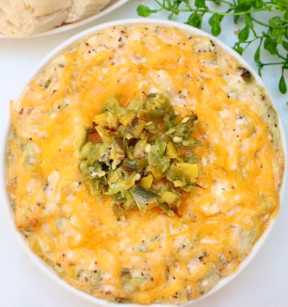 A bowl of baked cheesy dip topped with chopped green chilies sits on a white surface, with a sprig of greenery and a plate of crackers or bread in the background.