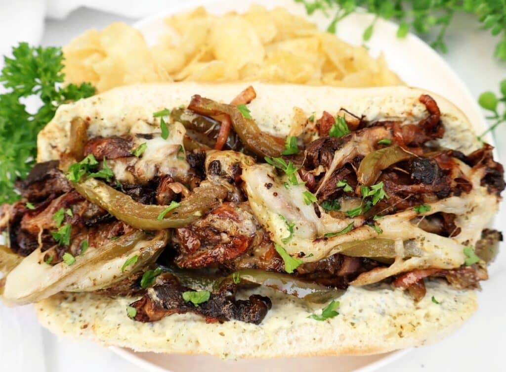 A loaded sandwich roll filled with grilled vegetables, melted cheese, and fresh herbs, served with potato chips on a white plate, garnished with parsley.