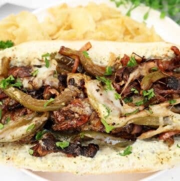 A loaded sandwich roll filled with grilled vegetables, melted cheese, and fresh herbs, served with potato chips on a white plate, garnished with parsley.