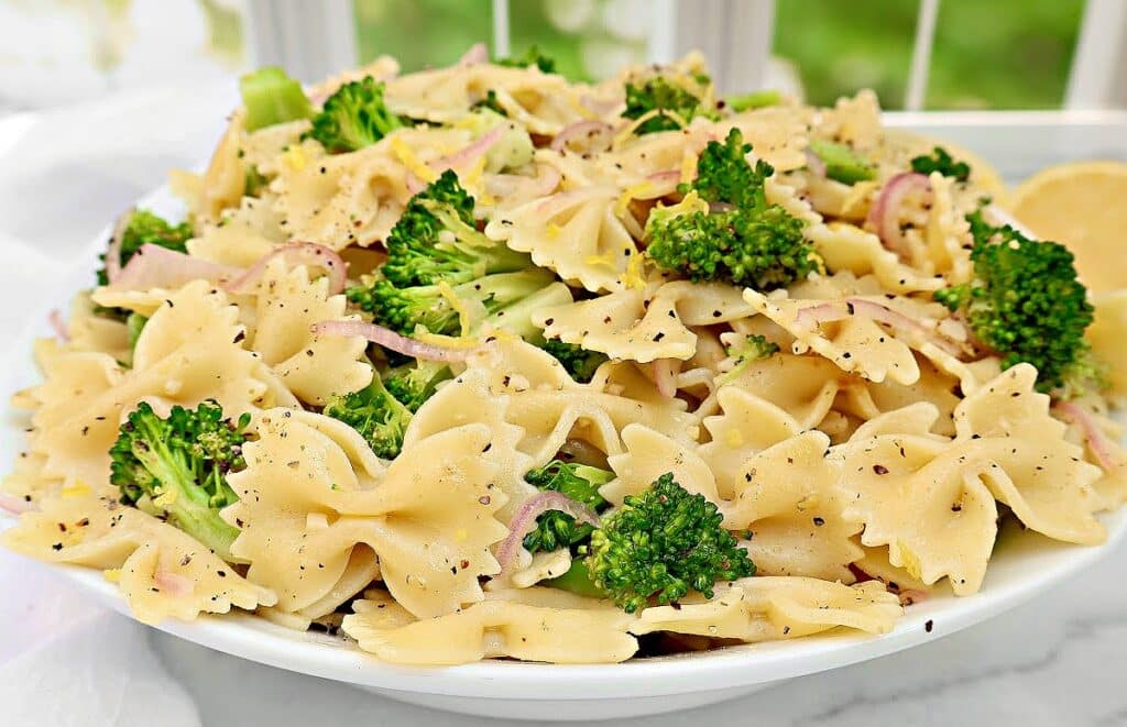 A white plate filled with bow-tie pasta, broccoli florets, and thinly sliced red onions, all seasoned with black pepper, sits on a white surface with a blurred green background.