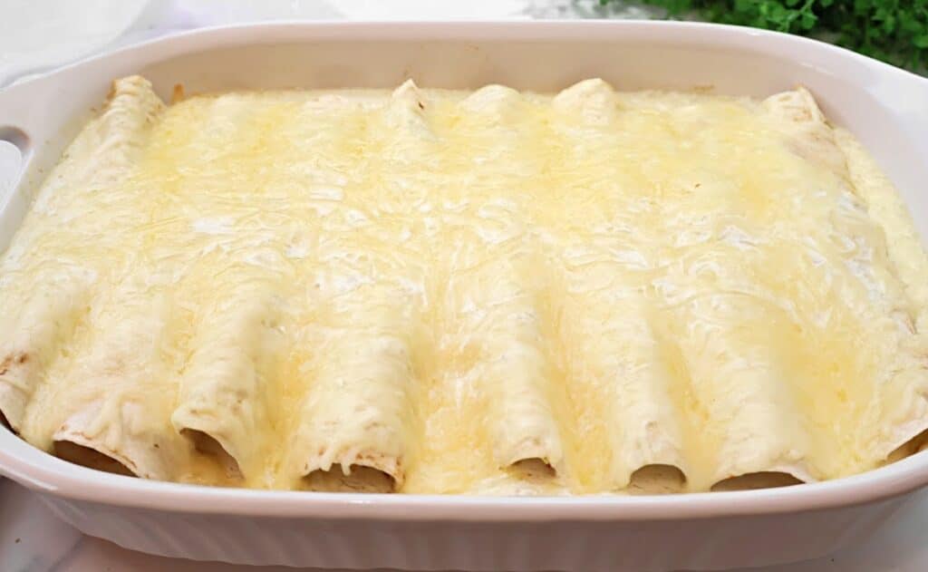 A white casserole dish filled with enchiladas covered in melted cheese and creamy sauce, baked until golden, shown from above.