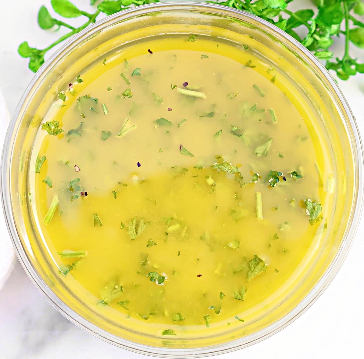 A clear bowl filled with yellow salad dressing, speckled with green herbs, sits on a white surface with fresh greens visible in the background.