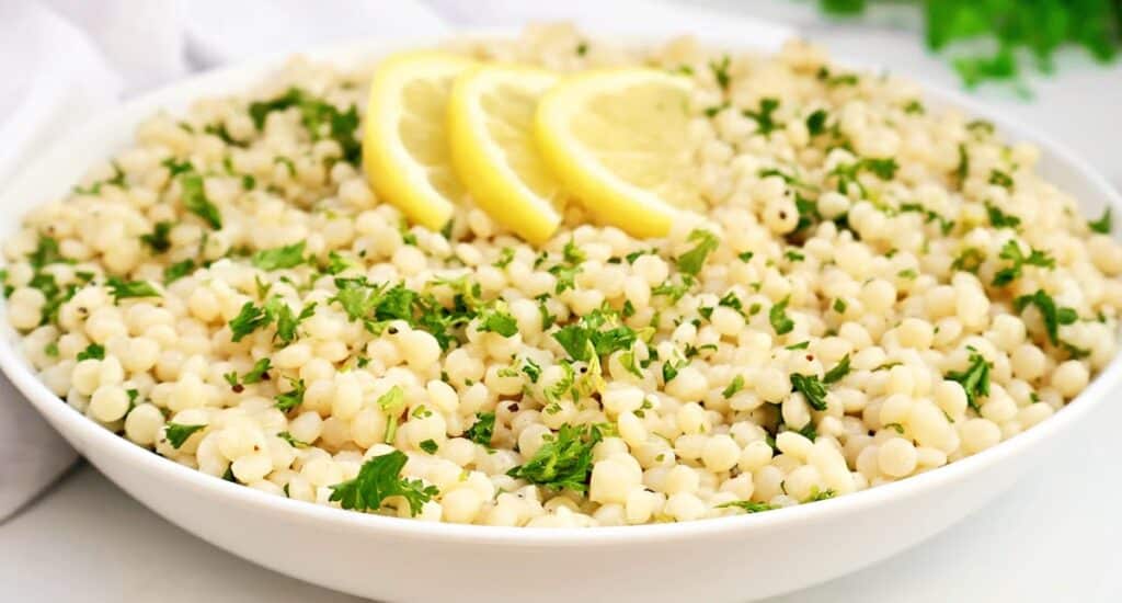 A white bowl filled with Israeli couscous garnished with chopped parsley and topped with three lemon slices, set on a white surface.