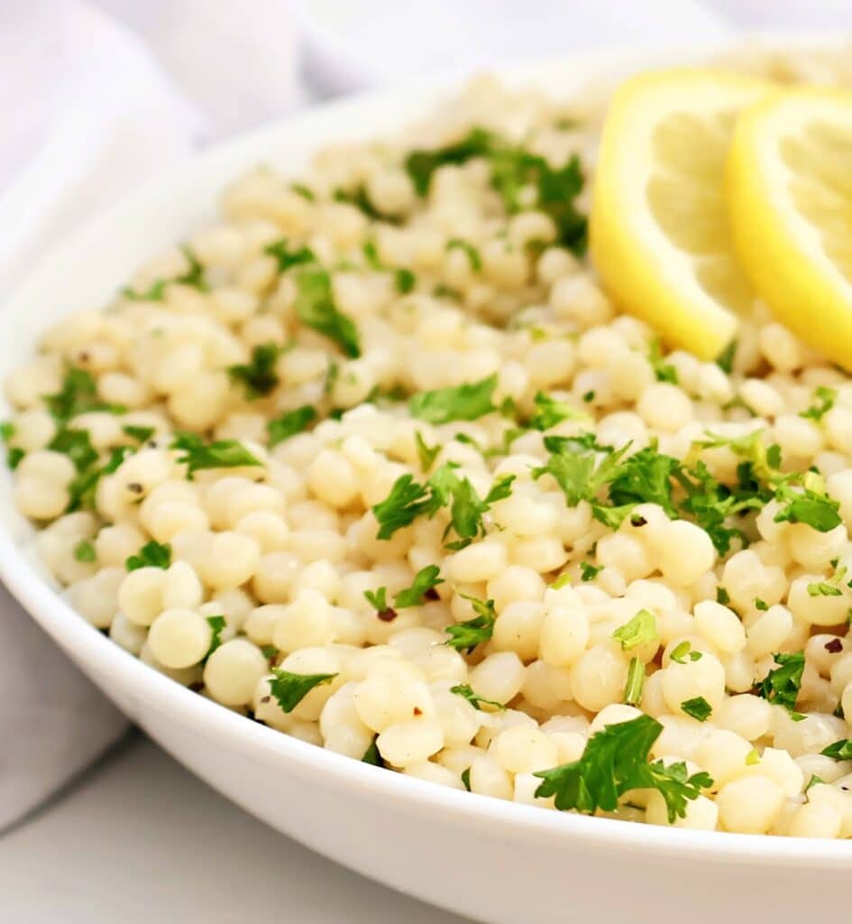 A white bowl filled with cooked pearl couscous, garnished with chopped parsley and two lemon slices.