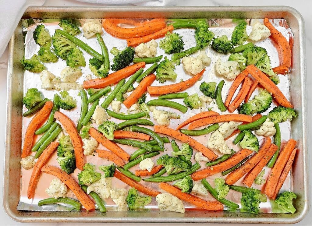A baking sheet lined with foil holds an assortment of raw vegetables, including broccoli florets, cauliflower, carrot sticks, and green beans, all seasoned and ready to be roasted.
