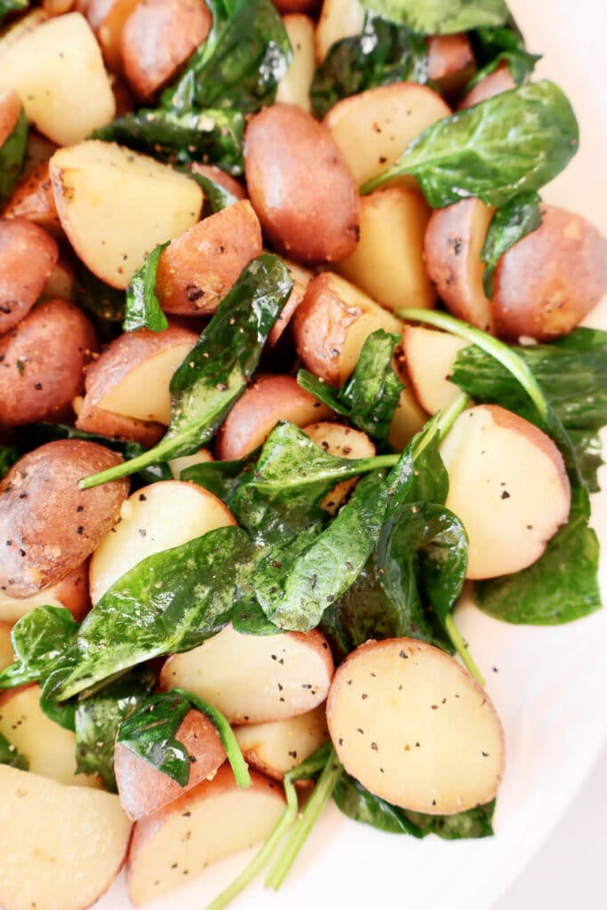 Halved red potatoes mixed with fresh spinach leaves, seasoned with black pepper, on a white plate. The dish appears light, healthy, and vibrant with green and golden colors.
