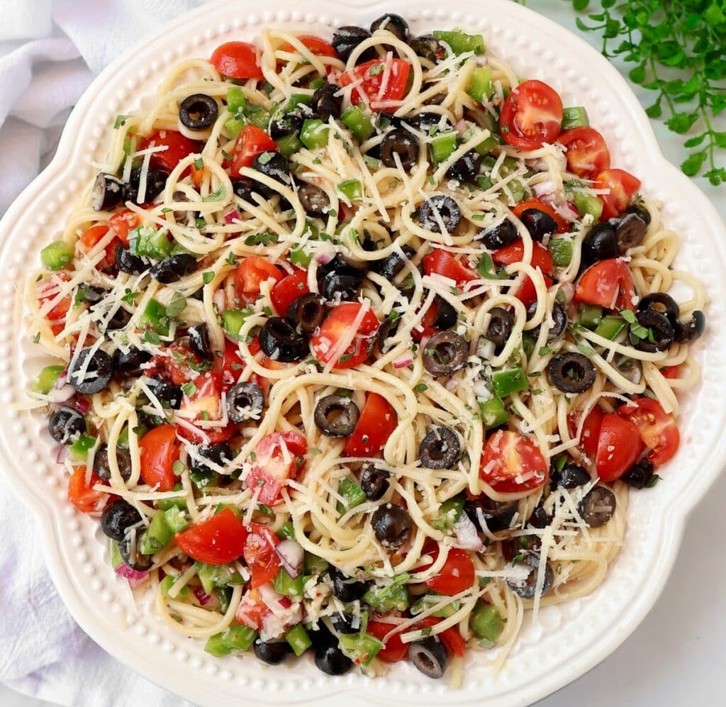 A white plate filled with spaghetti pasta salad, topped with black olives, cherry tomatoes, chopped green bell peppers, and shredded cheese. The colorful salad is presented on a textured white cloth.